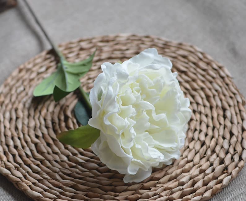 May include: A single white artificial peony flower with green leaves on a woven brown wicker mat.