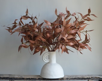 May include: A bouquet of dried eucalyptus branches in a white ceramic vase with a unique handle. The branches are a deep reddish-brown color.