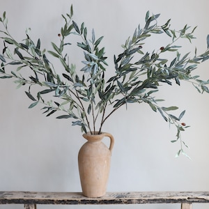 May include: A bouquet of artificial olive branches with green leaves and small brown olives in a light brown ceramic vase. The vase has a handle and is sitting on a rustic wooden table.