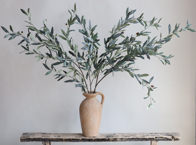 May include: A bouquet of artificial olive branches with green leaves and small brown olives in a light brown ceramic vase. The vase has a handle and is sitting on a rustic wooden table.