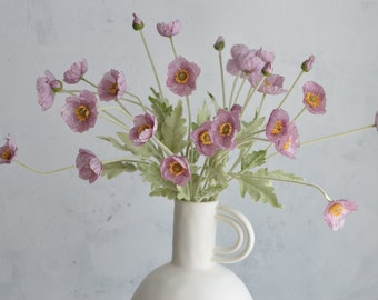 May include: A bouquet of pale pink silk poppy flowers with yellow centers arranged in a white ceramic vase with two handles.