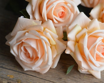 May include: A bouquet of four peach-colored artificial roses with a soft, velvety texture. The roses are arranged on a rustic wooden surface.