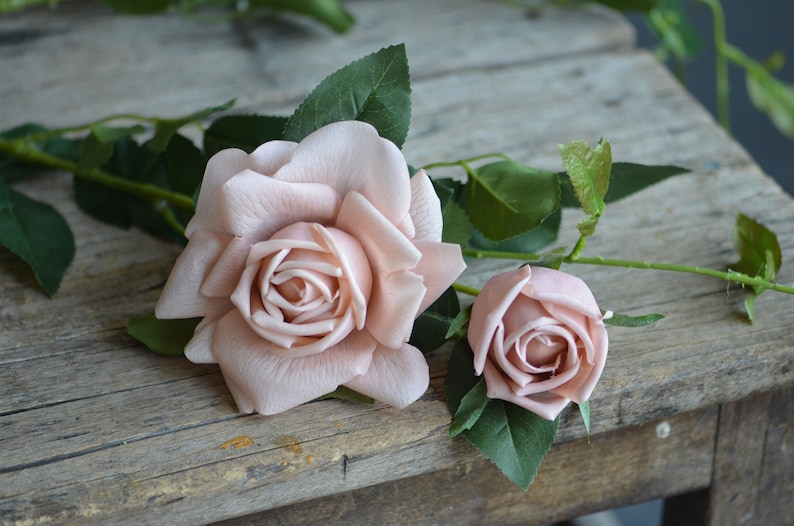 Op de afbeelding: Twee roze kunstbloemen met groene bladeren op een rustiek houten oppervlak.