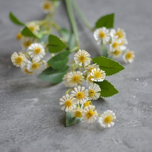 Puede incluir: Un ramo de flores artificiales blancas con centros amarillos y hojas verdes. Las flores están dispuestas en un racimo y se encuentran sobre un fondo gris.