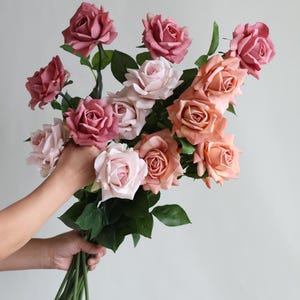 Puede incluir: Un ramo de rosas artificiales en tonos rosa y melocotón. Las rosas están dispuestas en un ramo sostenido a mano con tallos verdes.