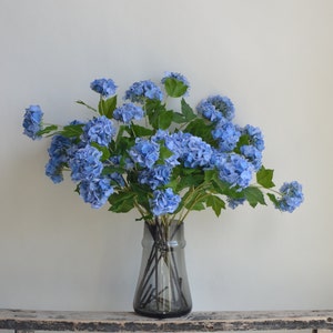 May include: A bouquet of blue hydrangeas in a clear glass vase with a textured surface. The vase is sitting on a weathered wooden shelf.