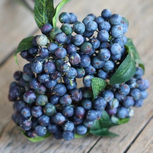May include: A close-up of a cluster of artificial blueberries with green leaves. The blueberries are a deep blue color with some lighter blue and purple hues. The leaves are a vibrant green. The arrangement sits on a wooden surface.