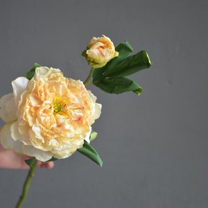 May include: A single artificial peach-colored peony flower with green leaves. The flower is in bloom and has a bud.