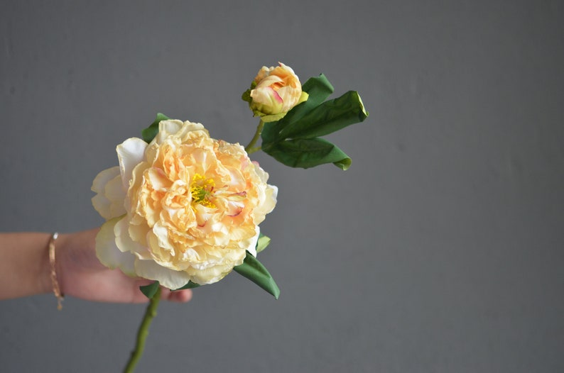 May include: A single artificial peach-colored peony flower with green leaves. The flower is in bloom and has a bud.