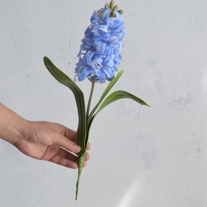 May include: A single artificial hyacinth flower with light blue petals and green leaves. The flower is held against a white background. The stem is a dark green color.