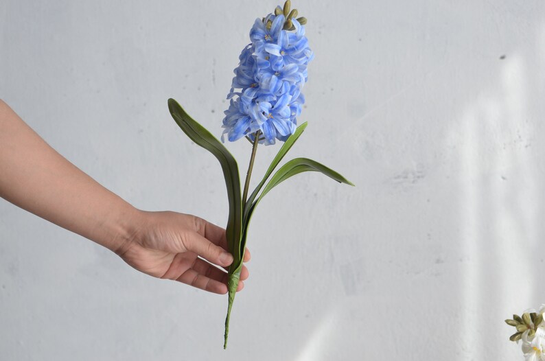 May include: A single artificial hyacinth flower with light blue petals and green leaves. The flower is held against a white background. The stem is a dark green color.