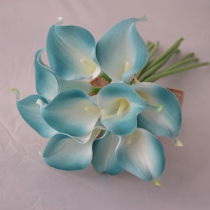 May include: A bouquet of artificial calla lilies in shades of teal and white. The flowers are clustered together and tied with a burlap ribbon. Green stems extend from the base of the bouquet, creating a natural look. Ideal for weddings or home decor.