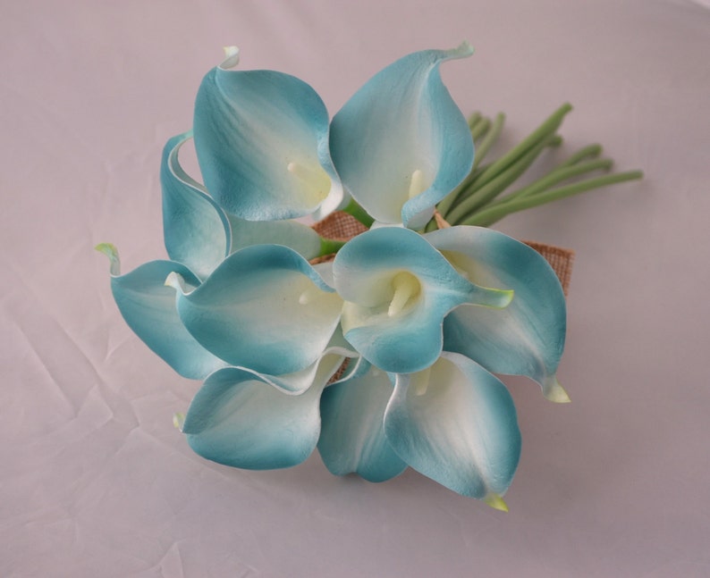 May include: A bouquet of artificial calla lilies in shades of teal and white. The flowers are clustered together and tied with a burlap ribbon. Green stems extend from the base of the bouquet, creating a natural look. Ideal for weddings or home decor.