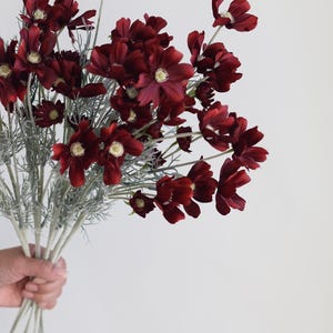 May include: A bouquet of burgundy artificial flowers with green stems. The flowers are arranged in a hand.