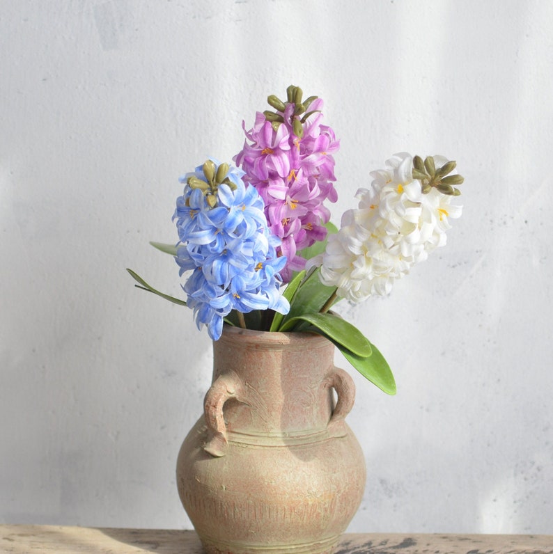 May include: A rustic, brown ceramic vase holds three hyacinth flower stems. The flowers are in shades of blue, purple, and white, with green leaves. The vase has two handles and sits on a wooden surface.
