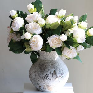 May include: A bouquet of white peonies with green leaves in a textured, gray vase. The flowers are in various stages of bloom, creating a full and lush arrangement. The vase has a rustic, weathered appearance.