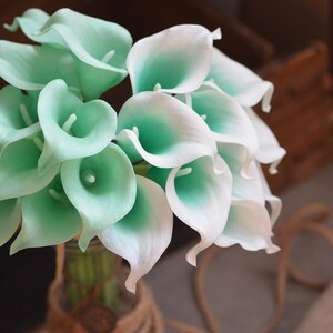 May include: A bouquet of artificial calla lilies in shades of white and light green. The flowers are arranged in a vase and are ready for a wedding or special event.