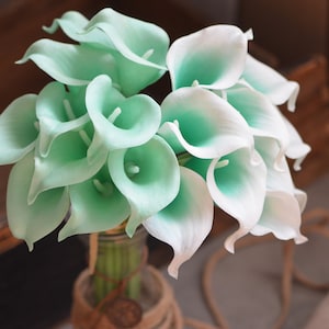May include: A bouquet of white and light green calla lilies arranged in a vase. The lilies are artificial and have a realistic appearance.