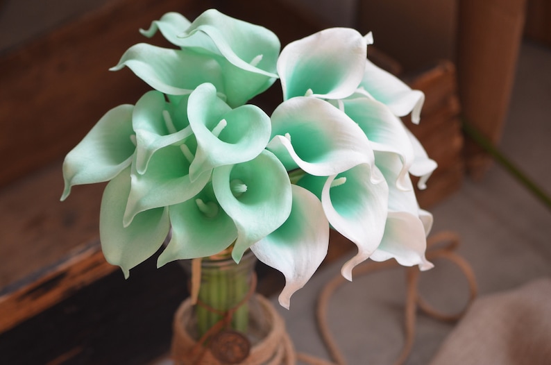 May include: A bouquet of white and light green calla lilies arranged in a vase. The lilies are artificial and have a realistic appearance.