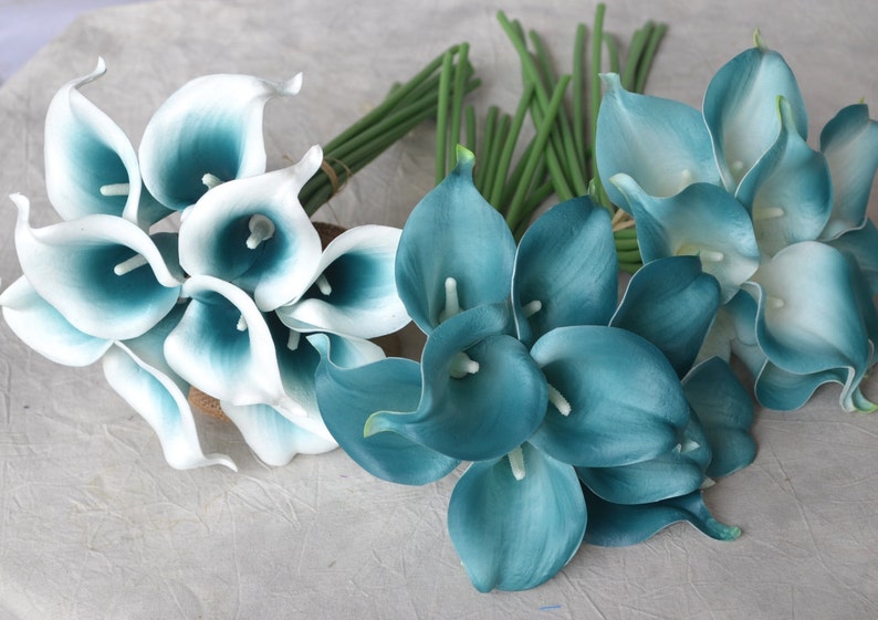 May include: Several bouquets of artificial calla lilies in shades of teal and white. The flowers have a smooth, waxy appearance and are arranged in bundles with green stems. The overall aesthetic is elegant and suitable for floral arrangements.