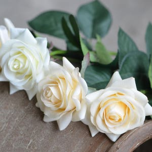 May include: Three cream-colored artificial roses with green leaves. The roses are arranged in a row on a brown surface.