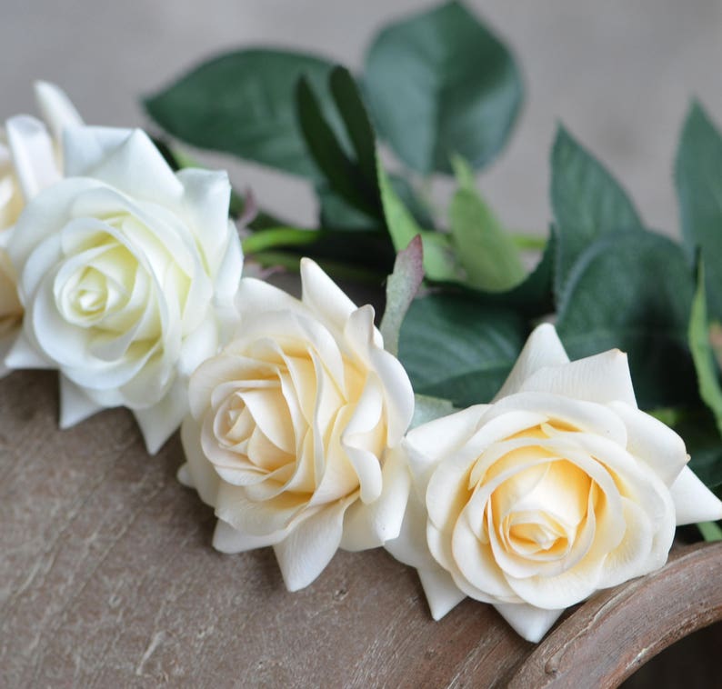 May include: Three cream-colored artificial roses with green leaves. The roses are arranged in a row on a brown surface.