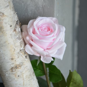 May include: A single pink artificial rose with green leaves and a brown stem. The rose is in focus and the background is blurred.