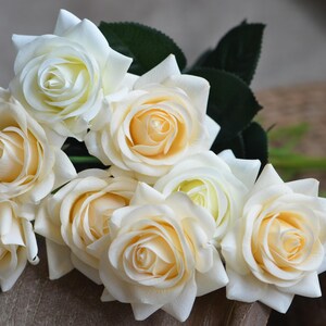 May include: A bouquet of cream-colored artificial roses with green leaves. The roses are arranged in a cluster, with the petals overlapping.