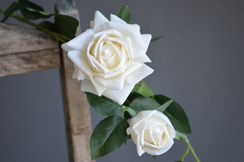 Op de afbeelding: Een enkele stengel met kunstmatige witte rozen met groene bladeren. De rozen staan in volle bloei en zien er realistisch uit.