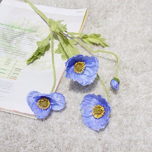 Puede incluir: Tres flores de amapola de seda azul con centros amarillos y tallos verdes. Las flores est&aacute;n dispuestas sobre una superficie blanca y peluda con un libro en el fondo.