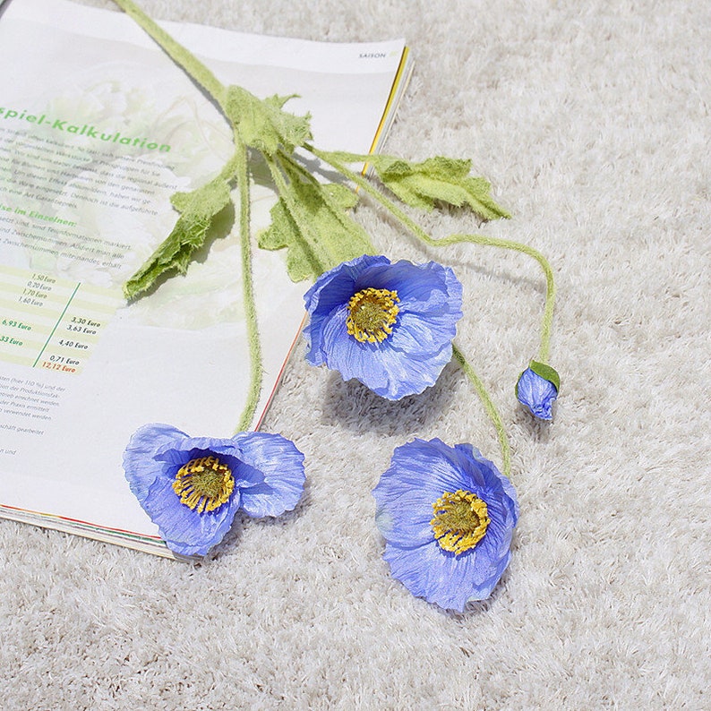 Puede incluir: Tres flores de amapola de seda azul con centros amarillos y tallos verdes. Las flores est&aacute;n dispuestas sobre una superficie blanca y peluda con un libro en el fondo.