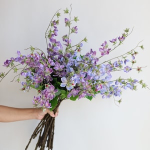 以下が含まれることがあります： 紫と青の色合いの造花の花束を人が持っています。花は繊細な花びらと緑の葉を持ち、茶色の茎があります。白い背景にアレンジされています。