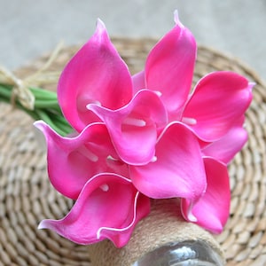 Puede incluir: Un ramo de calas rosas vibrantes con bordes blancos. Las flores están dispuestas con tallos verdes y atadas con hilo. El ramo se coloca sobre un fondo neutro, resaltando los pétalos de color rosa brillante.