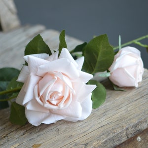 Op de afbeelding: Een enkele stengel kunstmatige rozen met twee lichtroze bloemen. De rozen zijn gerangschikt op een rustiek houten oppervlak.
