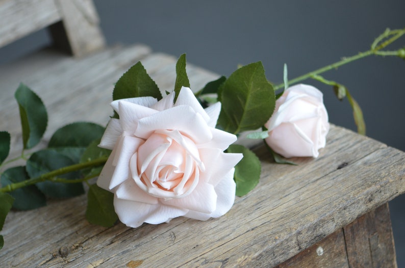 Op de afbeelding: Een enkele stengel kunstmatige rozen met twee lichtroze bloemen. De rozen zijn gerangschikt op een rustiek houten oppervlak.