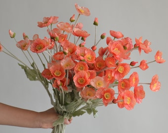 May include: A bouquet of coral-colored silk poppy flowers with green stems. The flowers are arranged in a hand-held bouquet.