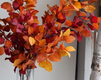 May include: A clear glass vase filled with branches of autumn leaves. The leaves display a vibrant array of colors, including shades of red, orange, and yellow. The arrangement is a decorative accent.