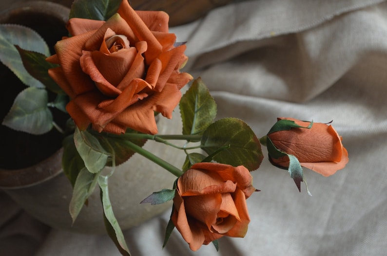 May include: A bouquet of three artificial roses in shades of orange and brown. The roses are arranged in a vase and have green leaves.