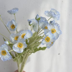 May include: A bouquet of light blue artificial poppy flowers with yellow centers. The flowers are arranged in a cluster and have a delicate, realistic appearance.