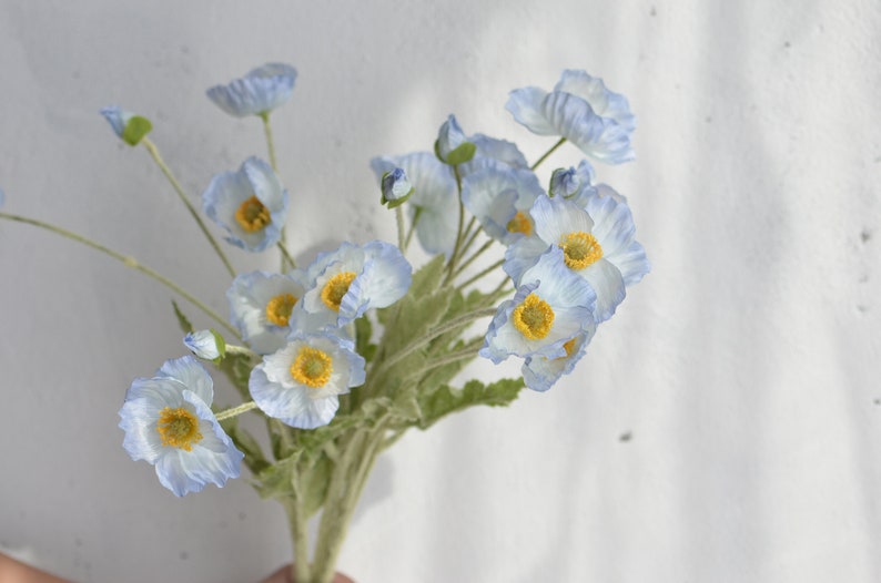 May include: A bouquet of light blue artificial poppy flowers with yellow centers. The flowers are arranged in a cluster and have a delicate, realistic appearance.
