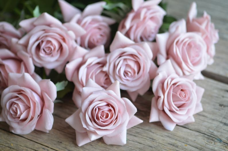 May include: A bouquet of blush pink artificial roses arranged on a wooden surface. The roses are made of fabric and have a realistic appearance.