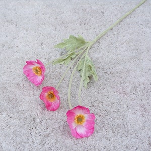 Puede incluir: Un ramo de tres flores artificiales rosas con tallos y hojas verdes. Las flores tienen un centro blanco y est&aacute;n dispuestas sobre un fondo blanco difuso.