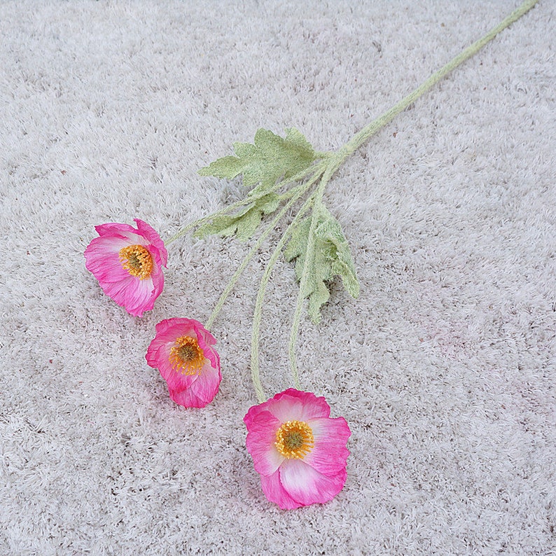 Puede incluir: Un ramo de tres flores artificiales rosas con tallos y hojas verdes. Las flores tienen un centro blanco y est&aacute;n dispuestas sobre un fondo blanco difuso.