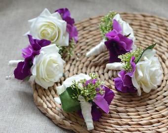 Puede incluir: Cinco rosas blancas con hortensias moradas y vegetación, dispuestas en un ramo. Los ramos están atados con una cinta blanca y se muestran sobre una superficie de mimbre tejido.