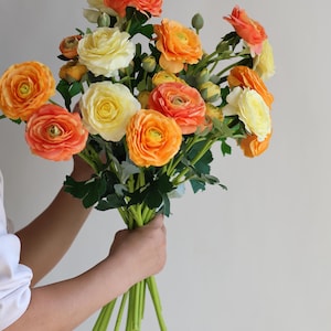 May include: A bouquet of artificial ranunculus flowers in shades of orange and yellow. The flowers have multiple layers of petals and are arranged in a hand-tied bouquet.