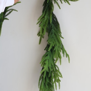 May include: A long, green artificial garland made of pine branches. The garland is hanging from a white wall.
