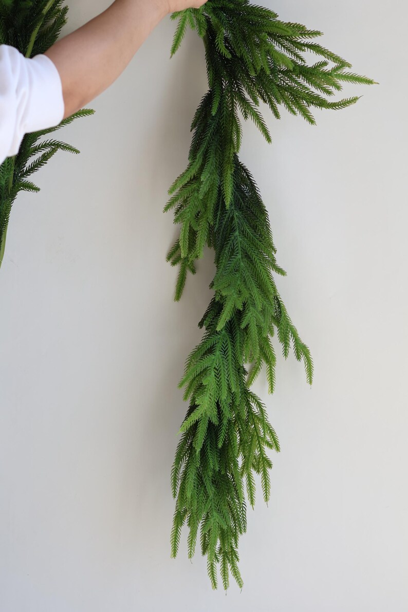 May include: A long, green artificial garland made of pine branches. The garland is hanging from a white wall.