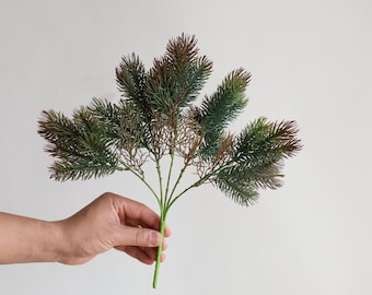 May include: A sprig of artificial greenery with green and brown needles. The stem is green, and the needles transition from green to brown at the tips. The sprig is held in a hand against a white background.