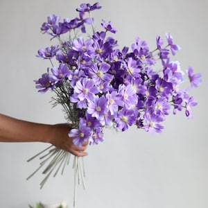 Könnte beinhalten: Ein Strauß künstlicher lila Blumen mit langen Stielen. Die Blumen sind in einem lockeren, natürlichen Stil angeordnet.
