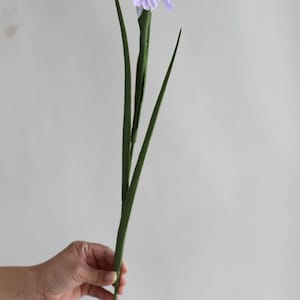 May include: A single artificial purple iris flower with green leaves. The flower is being held in a hand.